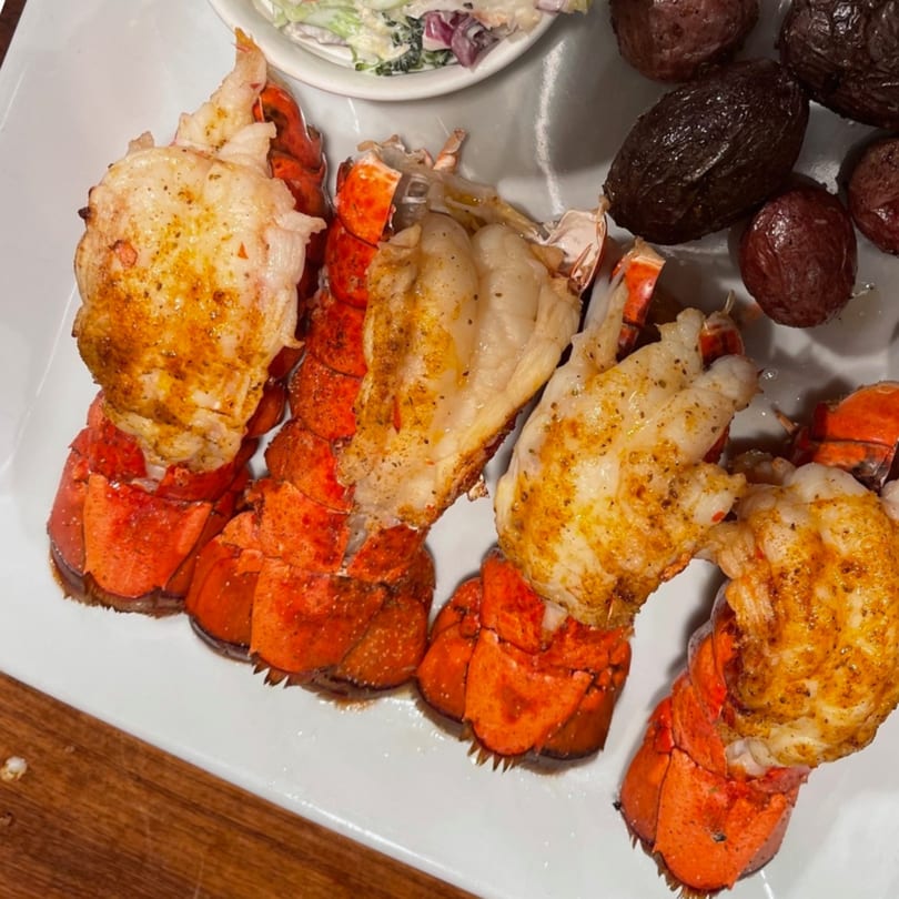 Four seasoned lobster tails with roasted potatoes and coleslaw on a white plate at Annie O's Kitchen.